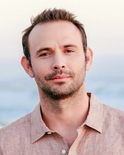 Alex Atkinson outdoors at the coast in soft afternoon light, in an open-necked linen shirt, the sea and a low horizon behind him.