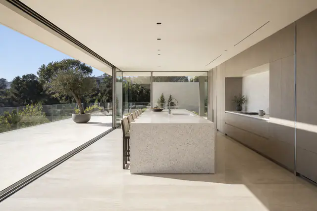 A Trousdale Estates kitchen in daylight: a terrazzo island under a flat plaster ceiling, oak cabinetry along the far wall, and a glass wall slid open to a travertine terrace and a potted olive.