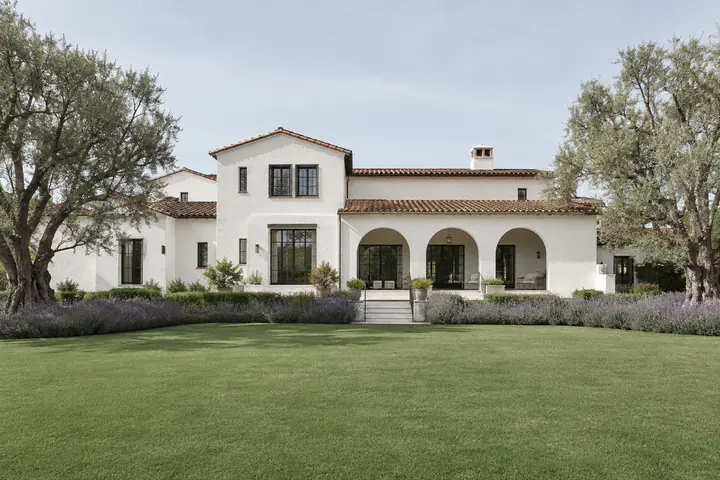 A white Spanish Colonial house in flat mid-morning light, its arched loggia and tiled roof behind a broad lawn edged with lavender, an olive tree at either side.