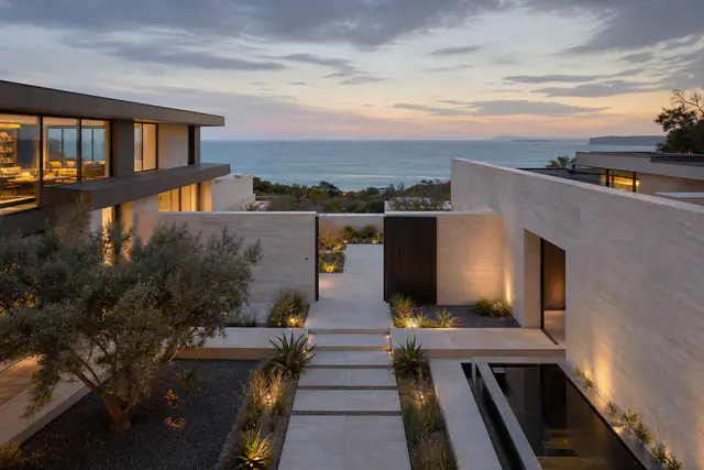 A courtyard house above Irvine Cove at dusk: two stone wings either side of a lit stone walkway, olive trees and low planting between them, and the ocean beyond the far wall.