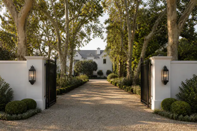 A white Spanish estate behind a gated drive in Brentwood, sycamores overhead in late light