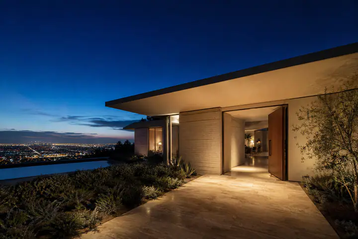 A Trousdale mid-century at blue hour, its front door standing open and lit, a travertine path leading to it and the city grid glowing far below.