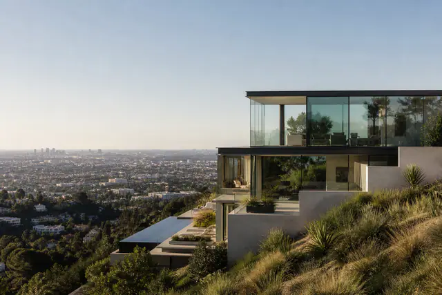 A glass-walled modern house cantilevered over the Hollywood Hills at golden hour, the city below