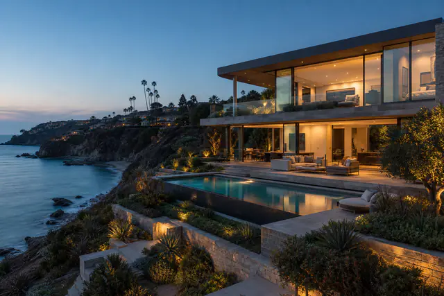 The flagship listing on the Hale & Cove sample site: a glass house stepping down a bluff at dusk, a lit pool on the terrace, a rocky cove and the open Pacific below.