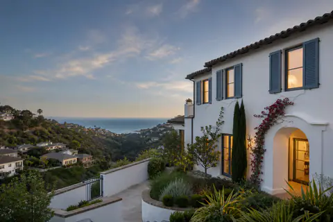 A Mediterranean villa on a hillside above the ocean at dusk, its shuttered windows lit, terraces and cypress stepping down towards the water.