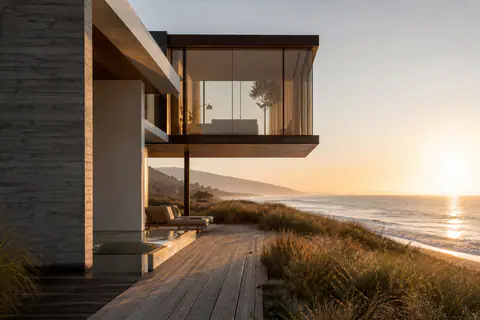 A glass room cantilevered over a bluff at golden hour, a teak deck running out through dune grass to a long empty beach below.