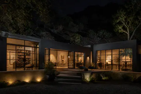 A low dark house on a canyon floor at night, its glass wall lit from within and a lit terrace of stone steps below a black hillside.